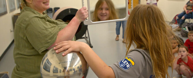 A girl looks in a mirror at her hair standing up with one hand on a van de graf generator