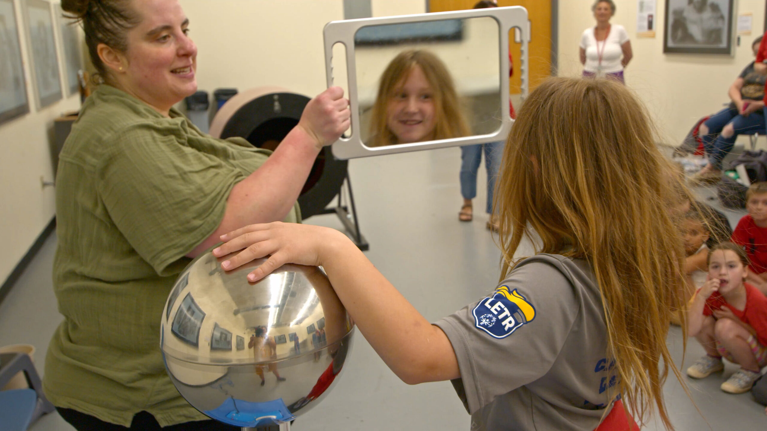 A girl looks in a mirror at her hair standing up with one hand on a van de graf generator