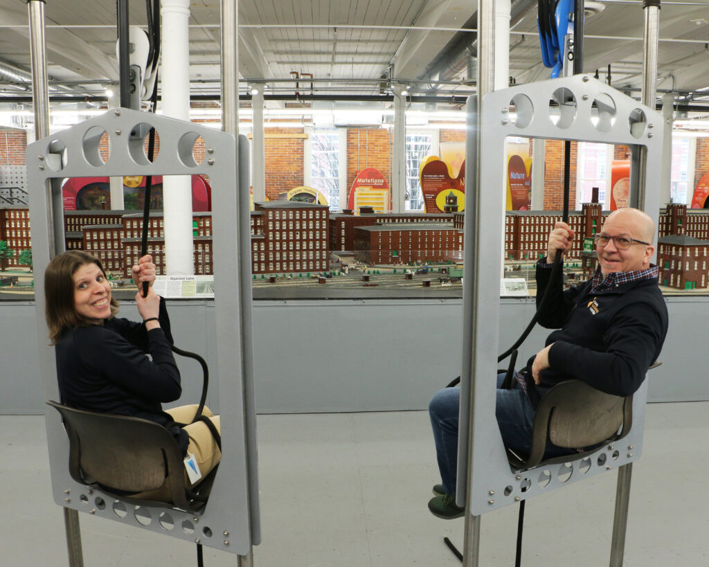 Two SEE employees sit on pulley chairs in front of the LEGO Millyard