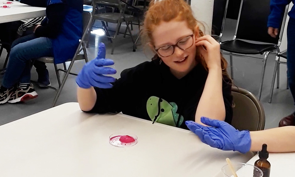a girl wearing gloves looks at a specimen in a petri dish