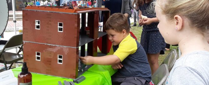 Children play with a LEGO building outdoors while a woman observes.