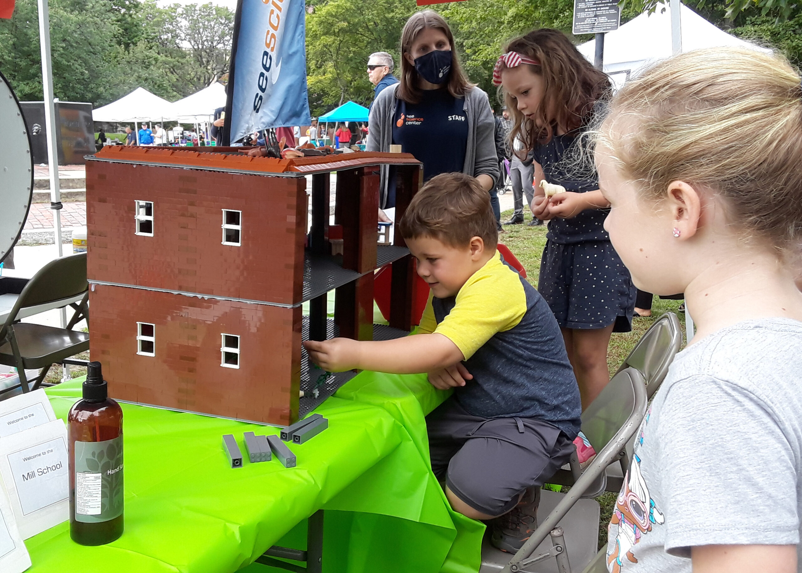 Children play with a LEGO building outdoors while a woman observes.
