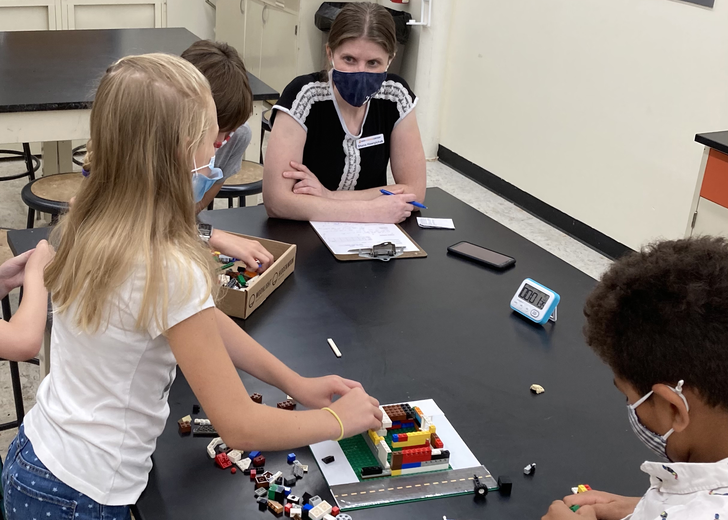 A woman with a clipboard observes children engaging in a tabletop exhibit.