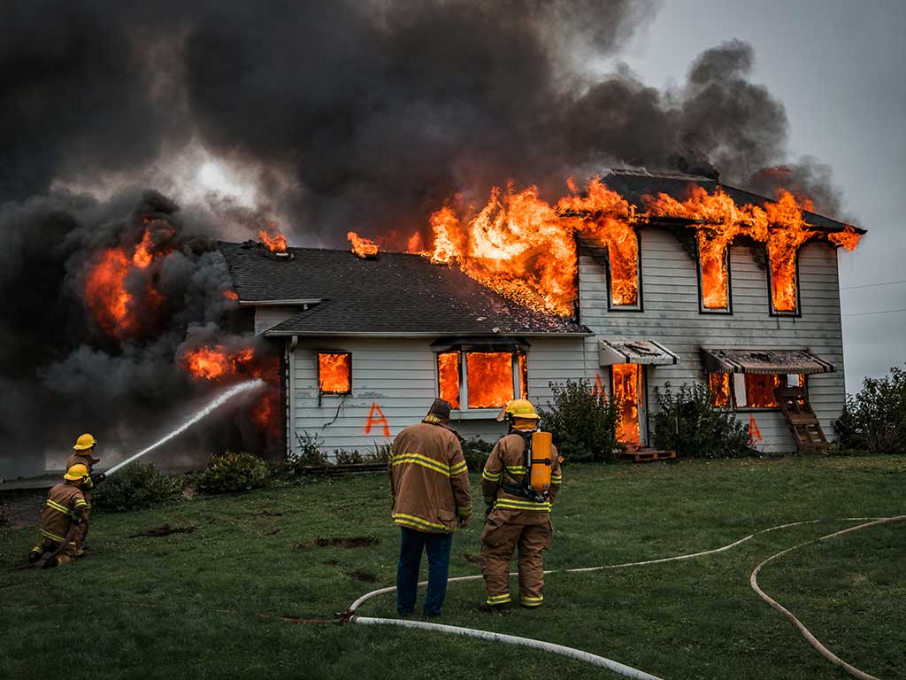 A firefighter sprays water on a building marked with an A as two other firefighters watch and take notes